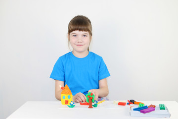 Smiling girl in blue molds house from plasticine on white table in white room