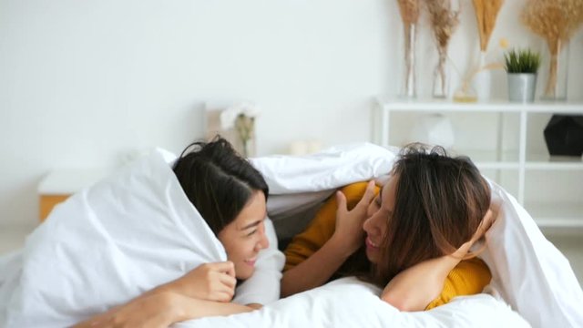 SLOW MOTION - Beautiful young asian women lesbian happy couple hugging and smiling while lying together in bed under blanket at home. Funny women after wake up. Lesbian couple together indoors concept