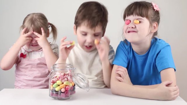 Three happy children play with candies near jar on table in white studio