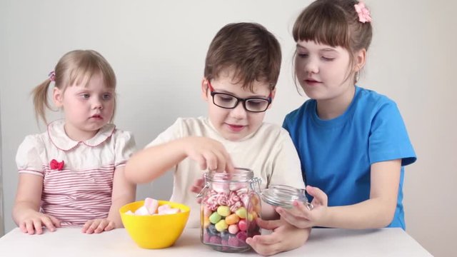 Three happy children take out marshmallows from jar and eat on table in white studio