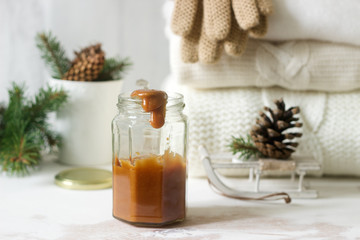 Fototapeta na wymiar Caramel in a glass jar on a winter background with a rug or pullover, gloves, a cup, spruce branches and cones.
