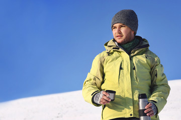 Man drinking hot coffee in thermos mug and looking into the mountains in snow, winter hike