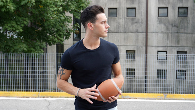 Handsome young man standing on street and holding American football ball.