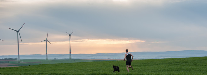 Obraz premium a man runs along the green field with a dog, with a view of the windmills