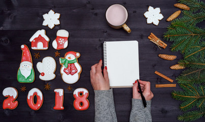 Christmas background with fir tree branches, cup of cocoa or chocolate, ginger cookies. Top view of blank notebook and woman hands with pen, free space. Girl writing to do list, copy space. Flat lay