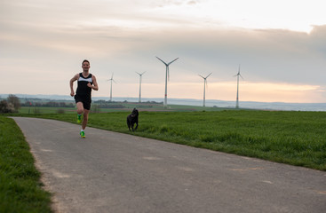 Obraz premium a man runs along the green field with a dog, with a view of the windmills