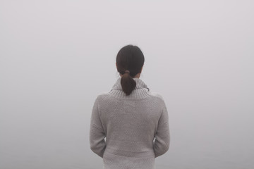 Junge traurige Frau steht mit dem Rücken zum Betrachter am Wasser Rückansicht Nebel