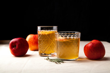 Two vintage glasses with apple cider on black background. Christmas beverages concept. Two red apples and rosemary sprig aside.  Warm backlight. Horizontal composition. Side view.