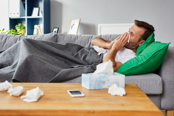 Sick man lying on sofa at home and blowing nose - obrazy, fototapety, plakaty