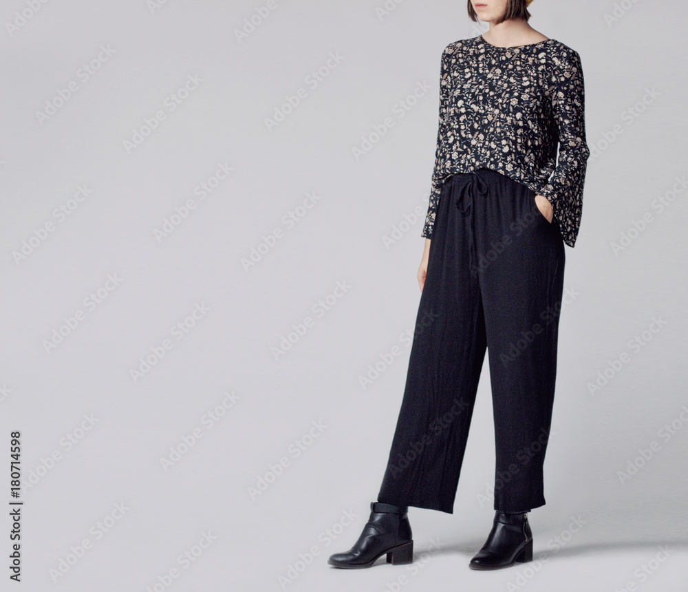 Wall mural Woman wearing stylish outfit with black patterned blouse, black high-waisted wide leg trousers and black ankle boots isolated on grey background. Copy space - Wall murals