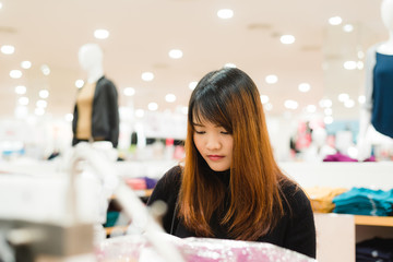 Half body shot of a happy asian young woman with shoulder bag looking at clothes hanging on the rail inside the clothing shop. Shopping, fashion, style and people woman concept.