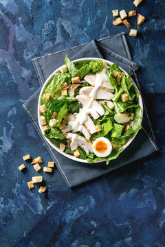 Classic Caesar salad with grilled chicken breast and half of egg in white ceramic plate. Served with croutons and dressing on textile napkin over dark blue texture background. Top view, space.