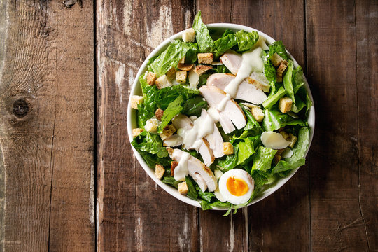 Classic Caesar salad with grilled chicken breast and half of egg in white ceramic plate. Served with croutons and dressing over old wooden background. Top view, space.
