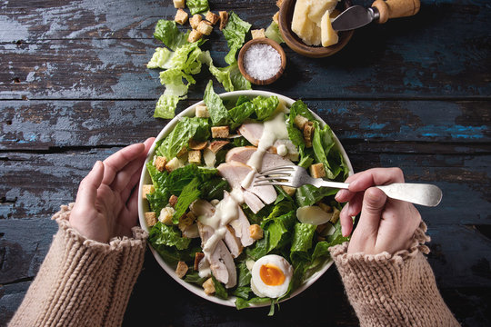 Female hands take by fork Classic Caesar salad with chicken breast in white ceramic plate. Served with ingredients above over old dark blue wooden background. Flat lay. Rustic style
