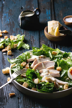 Classic Caesar salad with grilled chicken breast and half of egg in white ceramic plate. Served with fork and ingredients above over old dark blue wooden background. Rustic style