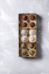 Variety of homemade dark chocolate truffles with cocoa powder, coconut, walnuts in golden gift box over gray texture background. Top view, copy space.