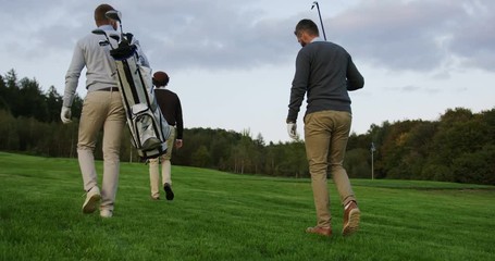 Three men with clubs and golf bag walking on the golf fied. Back view from below. Outside