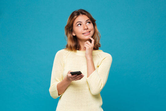 Pensive smiling woman in sweater holding smartphone and lookin up