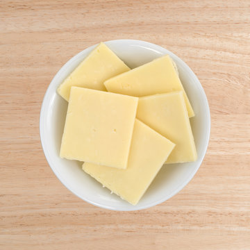Top view of a small white bowl filled with sharp cheddar cheese square slices on a wood table.