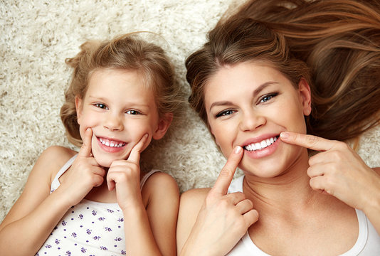 Portrait of beautiful daughter with mom showing their healthy white smile lying on carpet at home.