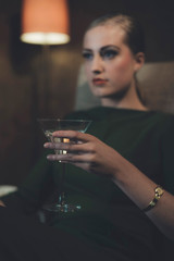 Retro 1960s woman sitting with cocktail glass in chair of hotel room at night.