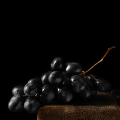 Black grapes on a wooden board on a dark background.  A minimalistic photograph in square format with copy space.
