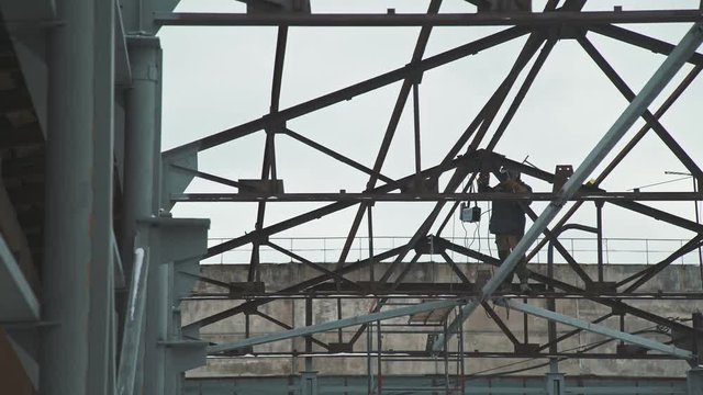 The welder in the protective helmet works at a height, welding the frame elements. Construction work: the creation of the roof of the premises, the metal frame of the building. Welder at work.