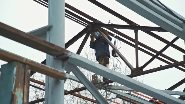 The welder in the protective helmet works at a height, welding the frame elements. Construction work: the creation of the roof of the premises, the metal frame of the building. Welder at work.