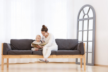 Pretty young mother reading a book to her little baby boy on a sofa in a light bedroom of house. Family holiday and togetherness.