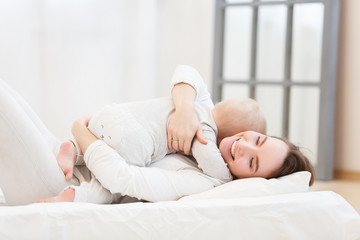 Obraz na płótnie Canvas Happy cheerful young mom smiling playing with her little baby boy lying in bed at home. family portrait, white clothes, light house interior