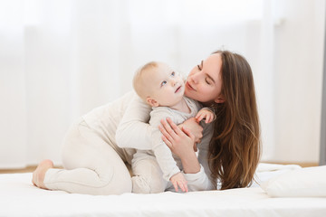 Happy cheerful young mom smiling playing with her little baby boy lying in bed at home. family portrait, white clothes, light house interior