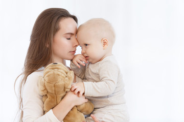 young caucasian mother holding her baby son in her arms before bedtime in a light bedroom. The family is dressed in white pajamas. Mother's Day. Mother's care and love