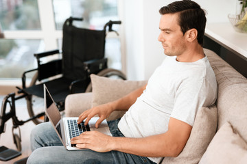 Fototapeta na wymiar The disabled person is sitting on the couch. He works for his laptop. next is his wheelchair. He sits on a large bright couch in his modern apartment.