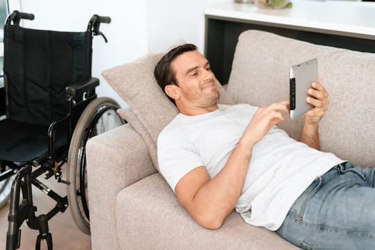 The disabled person lies on the couch and reads something on his tablet. Nearby is his wheelchair. He is on a large bright couch in his modern apartment.