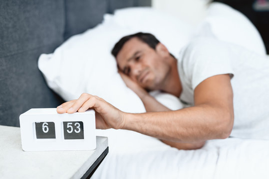 The disabled person sleeps in his white bed. There is an alarm clock in front of him. He frowns and pulls it off. He is in the bedroom of his modern apartment. The man is sleeping in a white T-shirt.