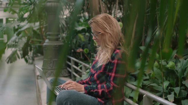 Soft focus shot of young woman in pleated shirt with long blonde hair, hipster or millennial generation work remotely on laptop while sit in park, freelance job ideas, unconventional office