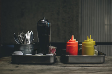 Close up image of bottles of mustard  ketchup  and salt on silver metal flavor tray in a burger shop, dark rustic wooden background with blank copy space