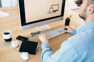 Close-up picture of a young man  working with a computer at his desktop.  He wears blue shirt. View from the side.