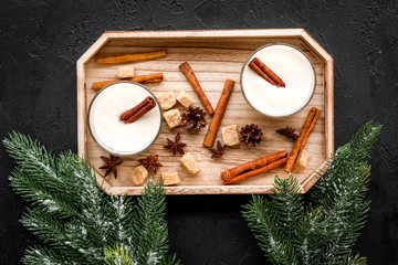 Homemade classic eggnog with cinamon and badian on wooden tray near spruce branch, pinecones on black background top view