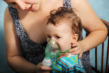Two year old baby girl inhaling from the inhaler, her mother holding her in the arms and comforting sick child. Treatment of a cough inhaler. A child in a mask for inhalations