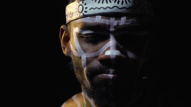 A bearded black man in white patterns and a ritual hat on his head, shakes his head to the beat of the music, slow motion