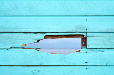 Bluish green painted fence of wood boards, with a hole through which you can see the surface of an ocean