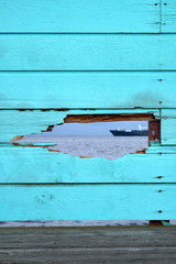 Bluish green painted fence of wood boards, with a hole through which you can see the surface of an ocean and a vessel sailing by