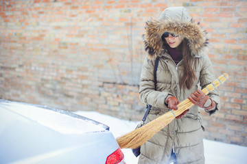 Happy girl cleaning her car from the snow in the city. By the house broom