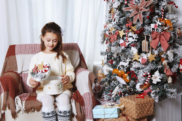 Beautiful girl sitting in a chair and opens a box with gift on background of Christmas tree