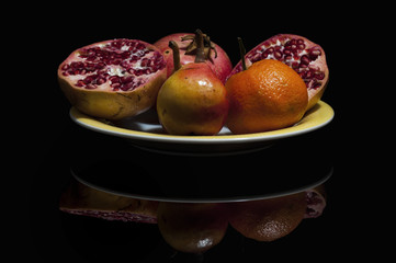 Italia, November  2017 - frutta autunnale still life in un piatto giallo su sfondo nero con riflesso. Pere, melograno, arance