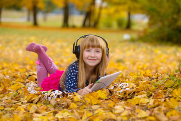 Little girl laying on grass and leaf and playing tablet pc with earphones