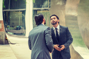 American Man with beard, dressing in gray suit, black tie, holding sunglasses, standing by metal mirror wall, looking at reflection. Concept of self assured, self esteem, self checking strategies..
