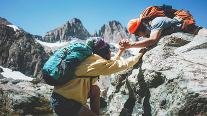 Traveler helping his friend to climb a rock. Young active couple with backpacks in stunning snowy...