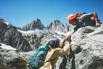 Traveler helping his friend to climb a rock. Young couple with backpacks in stunning snowy mountain wilderness near the lake